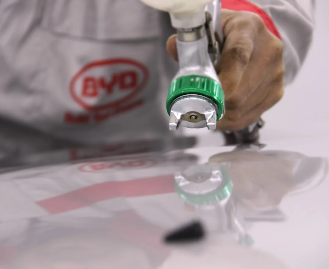 A garage painter in a respirator mask spraying a deep-red metallic basecoat onto a car door panel, with an HVLP spray gun, masking tape, and paint mixing cups on a workbench in the background